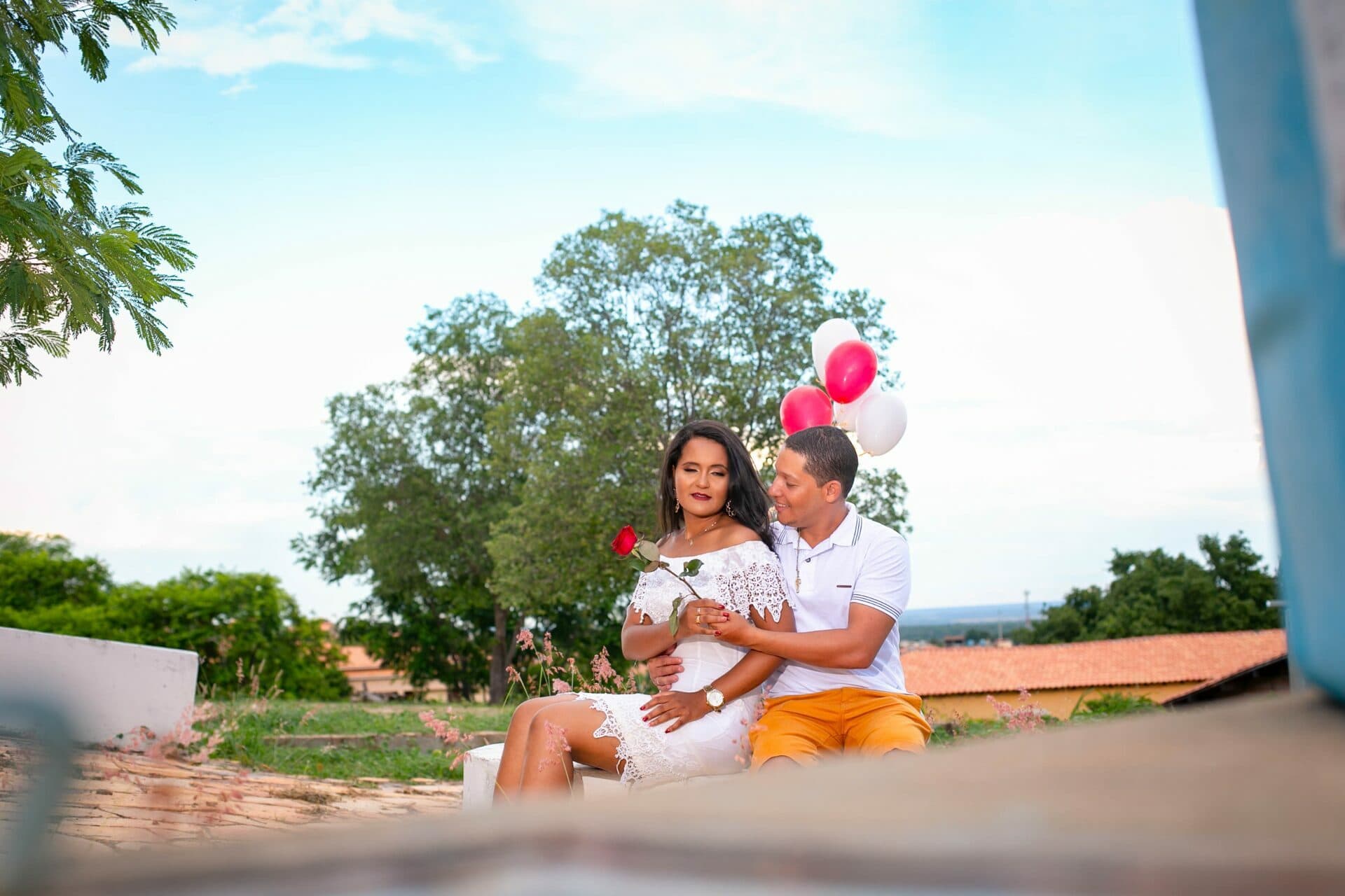 Casal sentado ao ar livre, com uma rosa e balões ao fundo.