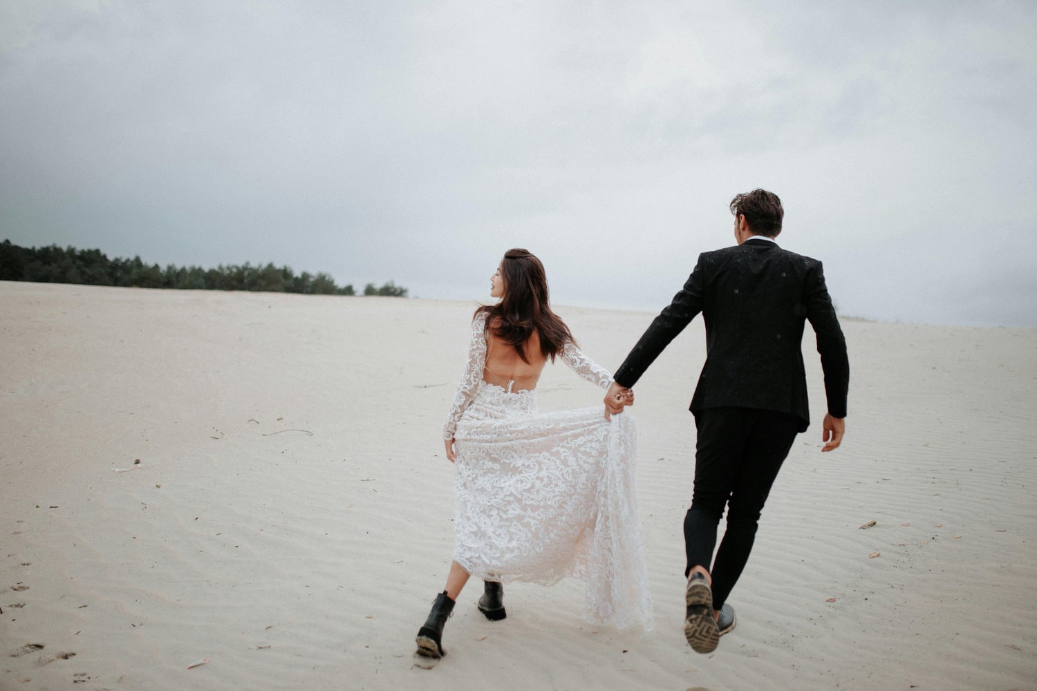 Casal de noivos caminha de mãos dadas entre dunas.